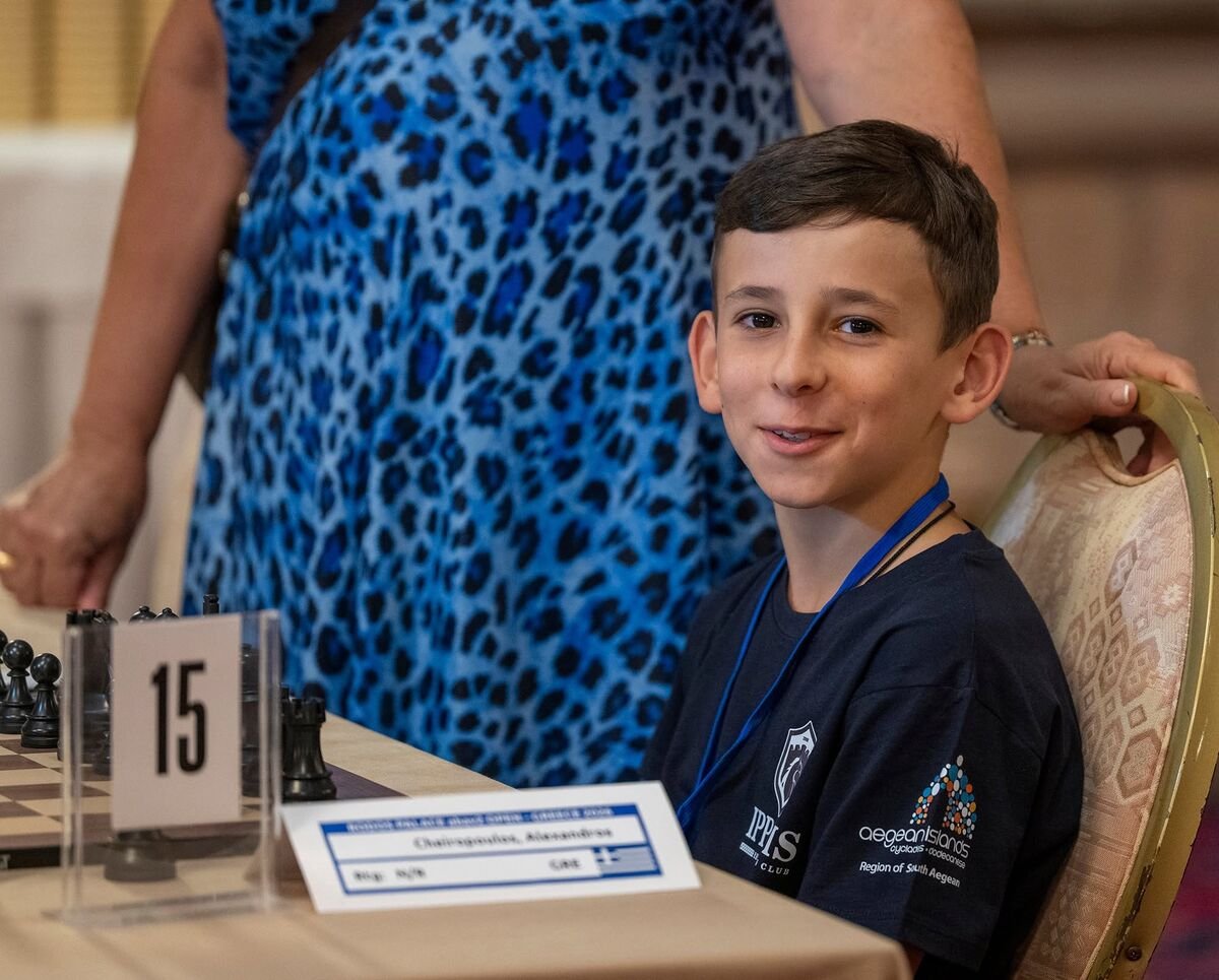 A boy wearing the Ippotis shirt smiles beside his chessboard