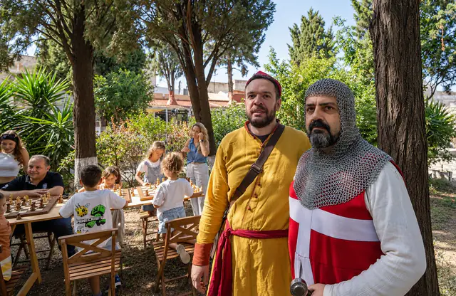 Δύο εκπρόσωποι του Ιππότη με φορεσιές εποχής μπροστά από τα τραπέζια με τις σκακιέρες.